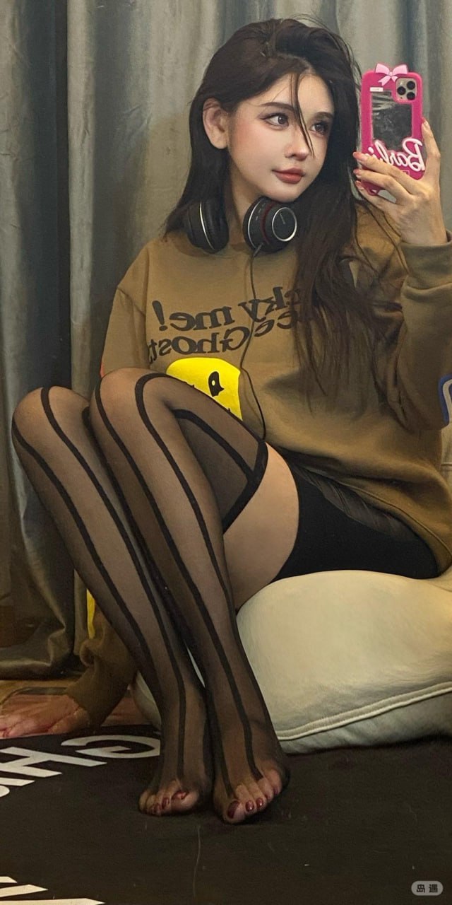 于芷晴穿棕色卫衣挂耳机在灰色窗帘前举手机